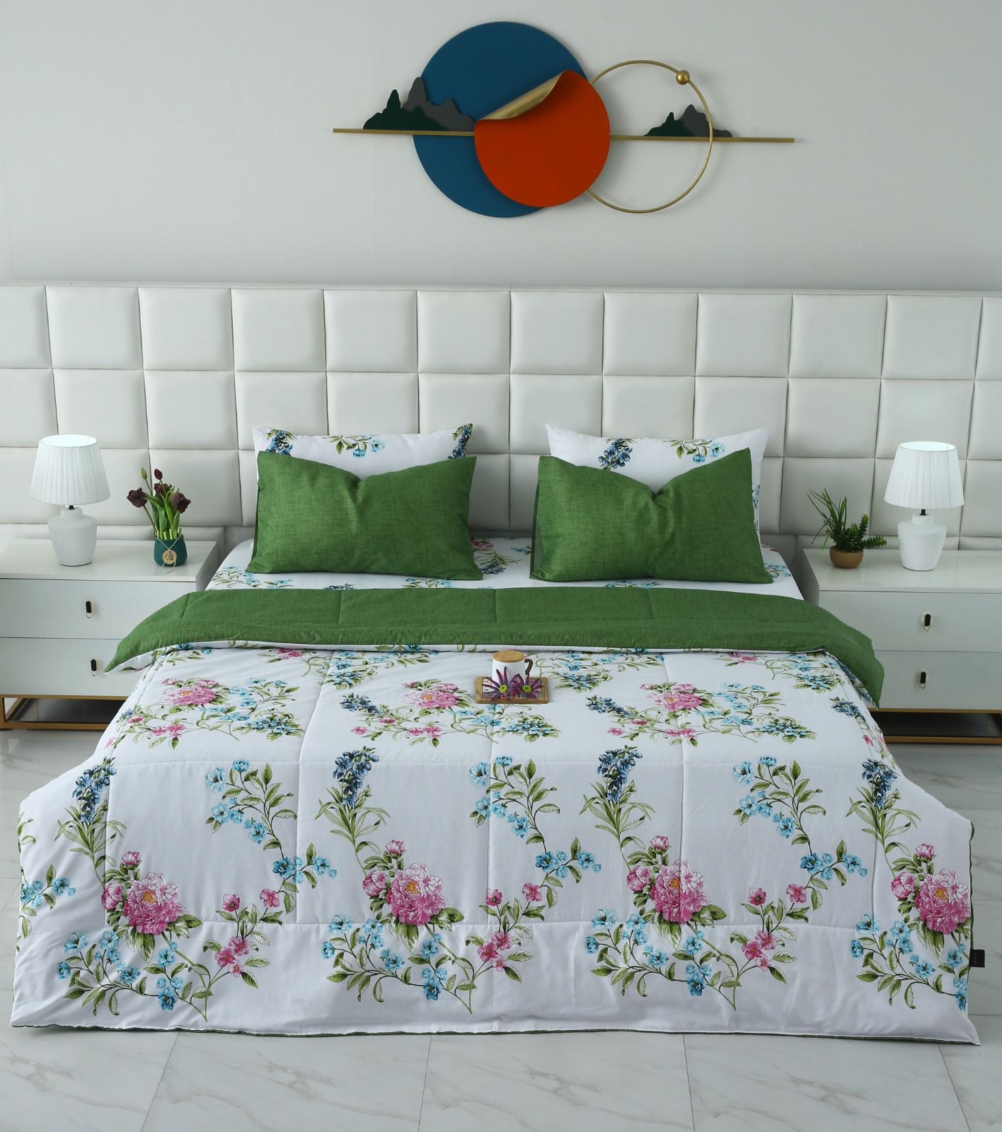 A neatly made bed in a bright bedroom setting, featuring a white comforter with a colorful floral print, two green pillows, and a green folded blanket. The background includes a white tufted headboard, white nightstands with lamps, and a decorative circular wall piece.