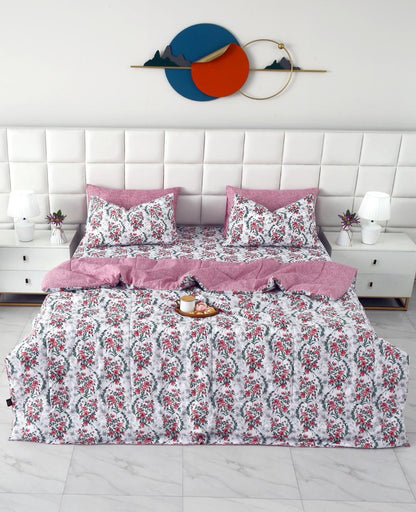 A neatly made bed in a modern bedroom setting, featuring a white bedding set with a pink and red floral pattern, two solid pink pillowcases, and a solid pink folded blanket. The background includes a white tufted headboard, white bedside tables with lamps, and a decorative wall piece.